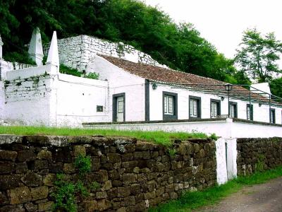 A long, single-story white building with a tiled roof and dark window frames, situated behind a low stone wall with a steep, vegetated hillside rising behind it.