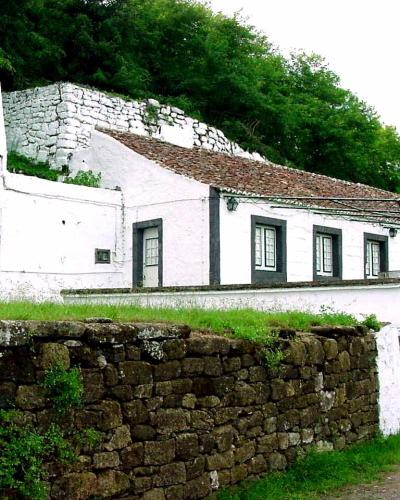 A long, single-story white building with a tiled roof and dark window frames, situated behind a low stone wall with a steep, vegetated hillside rising behind it.