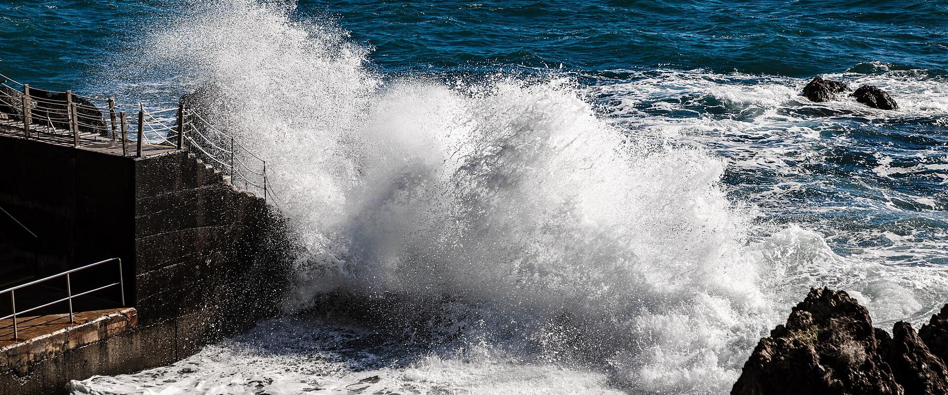 A large wave crashes violently against a dark concrete seawall and metal railing structure on a rocky coastline.