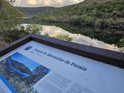 A close-up of an informational sign for "Parque de Merendas de Perais" with a view of a river and forested hills in the background.