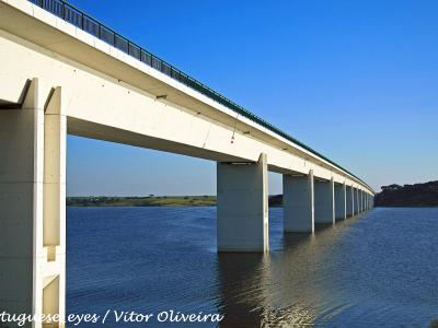 A long, white concrete bridge with tall pillars spanning across a large body of water under a clear blue sky, with a watermark reading "Portuguese eyes / Vitor Oliveira".