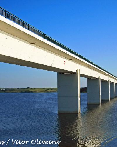 A long, white concrete bridge with tall pillars spanning across a large body of water under a clear blue sky, with a watermark reading "Portuguese eyes / Vitor Oliveira".