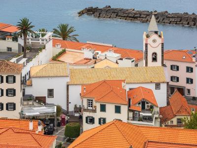 A high-angle view of the coastal town of Ponta do Sol, featuring white buildings with orange tiled roofs, a prominent church bell tower with a clock, and a stone breakwater in the ocean.