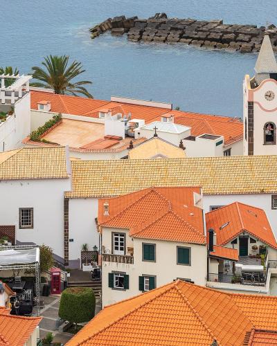 A high-angle view of the coastal town of Ponta do Sol, featuring white buildings with orange tiled roofs, a prominent church bell tower with a clock, and a stone breakwater in the ocean.