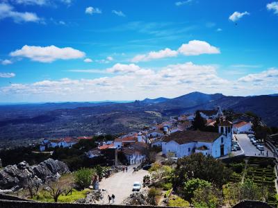 A high-angle view of the white-walled village of Marvão perched on a rocky hilltop, featuring a prominent church with a bell tower, terraced gardens, and a stone wall in the foreground, overlooking a vast landscape of hills and valleys under a blue s