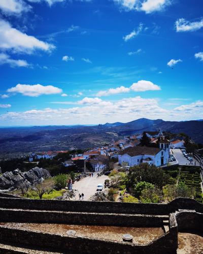 A high-angle view of the white-walled village of Marvão perched on a rocky hilltop, featuring a prominent church with a bell tower, terraced gardens, and a stone wall in the foreground, overlooking a vast landscape of hills and valleys under a blue s
