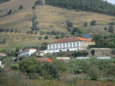 A white, two-story building with a terracotta-tiled roof sits on a hillside overlooking a valley, with an orange truck and industrial tanks visible in the foreground and terraced vineyards on the slope behind.