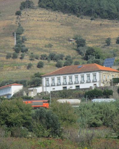 A white, two-story building with a terracotta-tiled roof sits on a hillside overlooking a valley, with an orange truck and industrial tanks visible in the foreground and terraced vineyards on the slope behind.