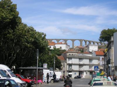 A street scene in a Portuguese town featuring a long, multi-arched stone aqueduct rising above white buildings with red-tiled roofs, with vehicles and pedestrians in the foreground.