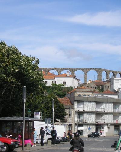 A street scene in a Portuguese town featuring a long, multi-arched stone aqueduct rising above white buildings with red-tiled roofs, with vehicles and pedestrians in the foreground.