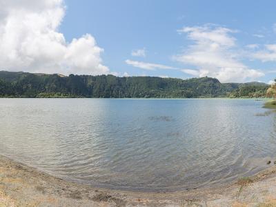 A panoramic view of a large, calm lake surrounded by green, rolling hills and a grassy shoreline under a partly cloudy sky.