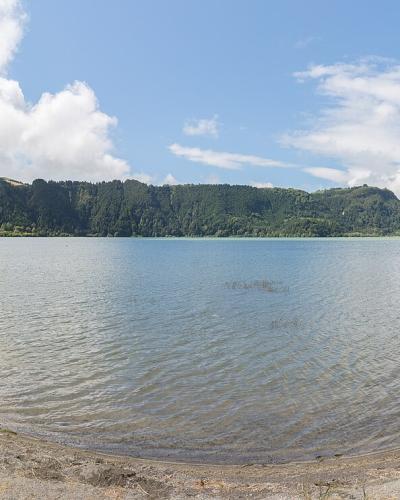 A panoramic view of a large, calm lake surrounded by green, rolling hills and a grassy shoreline under a partly cloudy sky.