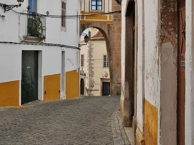A narrow cobblestone street in a historic town, featuring white buildings with yellow trim and a stone archway topped by a small chapel or bell tower structure.