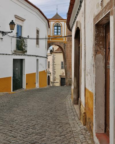 A narrow cobblestone street in a historic town, featuring white buildings with yellow trim and a stone archway topped by a small chapel or bell tower structure.