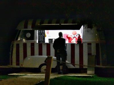 A vintage white van with red vertical stripes and a striped awning, modified into an ice cream serving window at night, with a customer standing in front and a menu board visible inside.