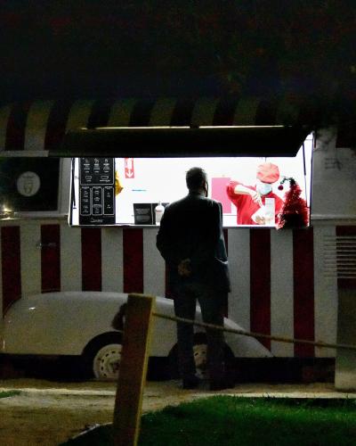 A vintage white van with red vertical stripes and a striped awning, modified into an ice cream serving window at night, with a customer standing in front and a menu board visible inside.