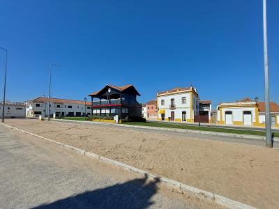 A wide shot of a sandy arena or parade ground bordered by a row of buildings, including a distinctive black wooden structure with a balcony and a white building with yellow trim.