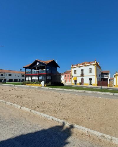 A wide shot of a sandy arena or parade ground bordered by a row of buildings, including a distinctive black wooden structure with a balcony and a white building with yellow trim.