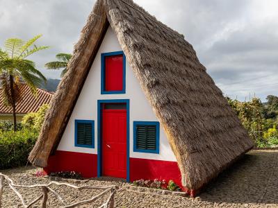 A traditional triangular A-frame house with a thatched roof, white walls, red base, and blue trim around the door and windows, situated on a cobblestone path with a wooden fence and vegetation.