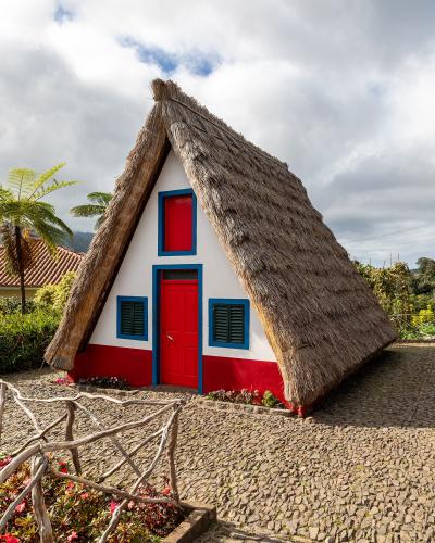 A traditional triangular A-frame house with a thatched roof, white walls, red base, and blue trim around the door and windows, situated on a cobblestone path with a wooden fence and vegetation.