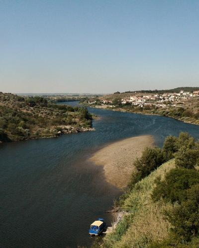 A wide river flows through a hilly, semi-arid landscape with a small village on the far bank and a blue tour boat docked near a sandy shoal; the photo is taken from an elevated stone vantage point, likely a castle wall.