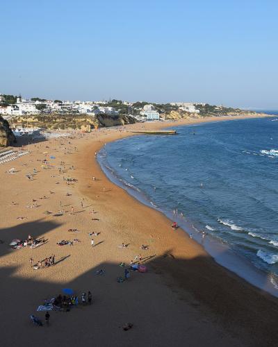 A high-angle view of a wide, sandy beach (Praia da Oura) with a large white hotel on the left, a line of beach umbrellas, and the Atlantic Ocean to the right.