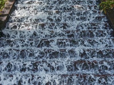 A close-up view of a man-made waterfall feature with water cascading down a series of dark, stepped stone tiers, flanked by lush green ferns and tropical foliage.