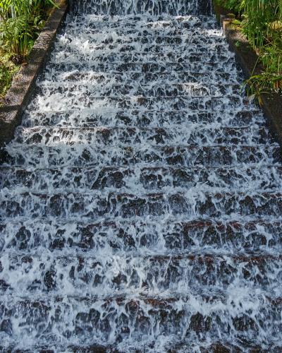 A close-up view of a man-made waterfall feature with water cascading down a series of dark, stepped stone tiers, flanked by lush green ferns and tropical foliage.