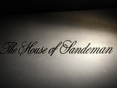 A close-up shot of a wall with the text "The House of Sandeman" in black cursive lettering, illuminated by a spotlight.