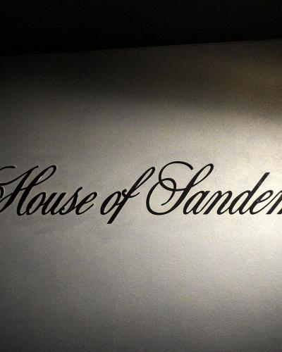 A close-up shot of a wall with the text "The House of Sandeman" in black cursive lettering, illuminated by a spotlight.