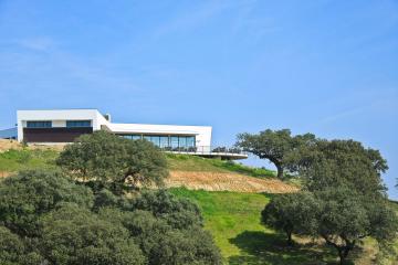 A modern white building with large glass windows and a terrace sits atop a grassy hill dotted with cork oak trees under a clear blue sky.