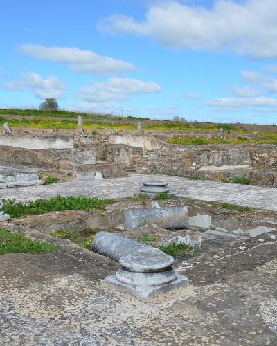 The image shows the archaeological remains of a Roman villa featuring stone foundations, column bases, and mosaic floor fragments under a blue sky.