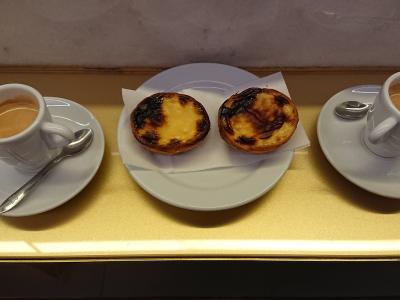 Two pastéis de nata on a white plate flanked by two cups of coffee on a gold-colored counter.