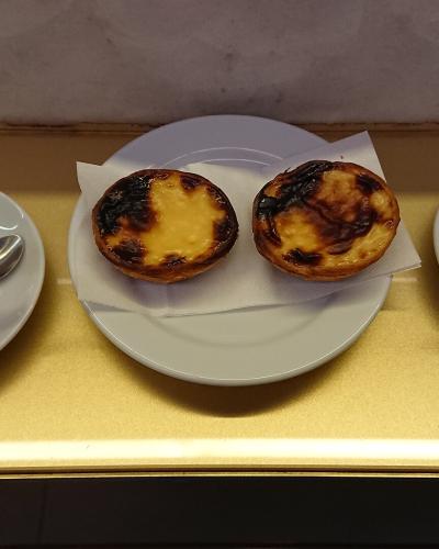 Two pastéis de nata on a white plate flanked by two cups of coffee on a gold-colored counter.