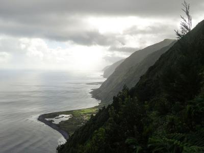 A high-angle landscape view of a dramatic, green, mountainous coastline curving along the ocean under a cloudy sky, with a small coastal settlement and a black sand beach visible in the valley below; the filename "Norte Pequeno after the rain (219