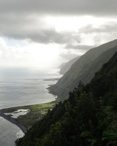 A high-angle landscape view of a dramatic, green, mountainous coastline curving along the ocean under a cloudy sky, with a small coastal settlement and a black sand beach visible in the valley below; the filename "Norte Pequeno after the rain (219