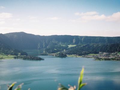 A scenic landscape photograph showing a large volcanic crater lake with two distinct bodies of water, surrounded by green hills and a small settlement on the shore.
