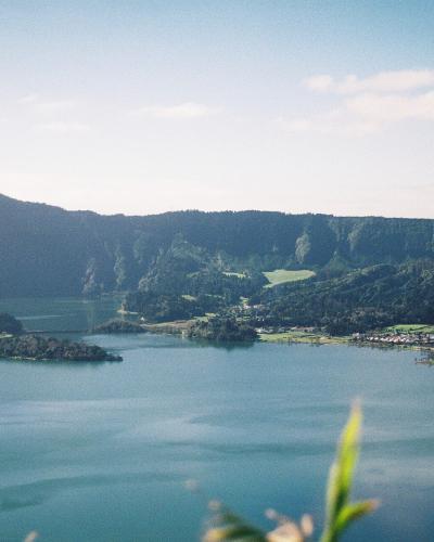 A scenic landscape photograph showing a large volcanic crater lake with two distinct bodies of water, surrounded by green hills and a small settlement on the shore.