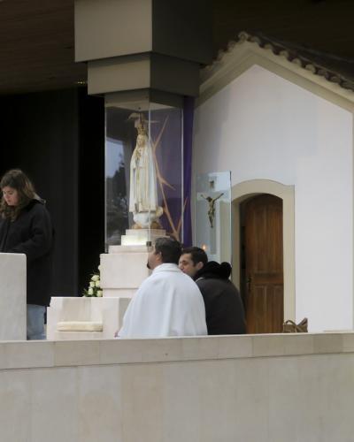 A small, white-walled chapel with a tiled roof and a blue tile mural of Our Lady of Fátima on the exterior wall, situated inside a larger modern building with a congregation seated in the foreground.