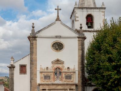 A white-walled church with a prominent stone portal, a statue of a saint in a niche above the door, and a tall bell tower with a conical spire, set against a cloudy sky.