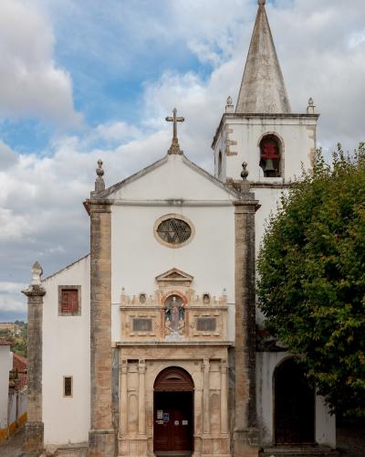 A white-walled church with a prominent stone portal, a statue of a saint in a niche above the door, and a tall bell tower with a conical spire, set against a cloudy sky.