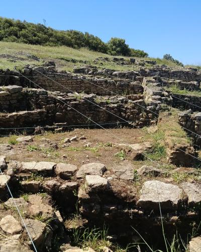 The image shows the stone foundations and excavated ruins of an ancient settlement on a grassy hillside, marked by thin white strings used for archaeological grid mapping.