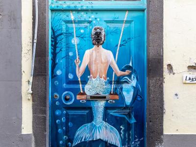 A close-up photograph of a blue wooden door painted with a mermaid on a swing, featuring the house number 105 above the frame.