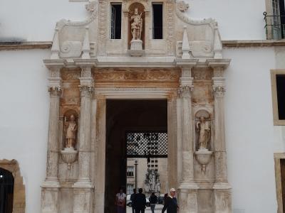 A large, ornate stone gateway with multiple statues, including a central figure of a crowned saint or king in a niche above the arch and flanking figures in side niches, set into a white building facade with people walking through the archway.