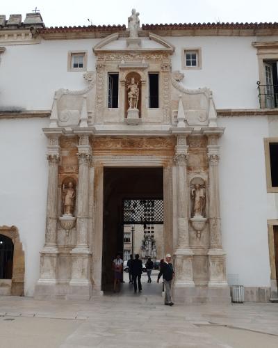 A large, ornate stone gateway with multiple statues, including a central figure of a crowned saint or king in a niche above the arch and flanking figures in side niches, set into a white building facade with people walking through the archway.