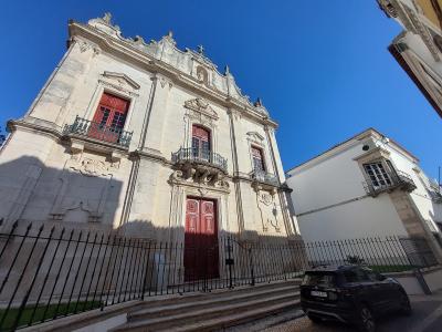 A low-angle photograph of a white stone church facade with red wooden doors and window shutters, featuring a black metal fence in the foreground and a black car parked on the street to the right.