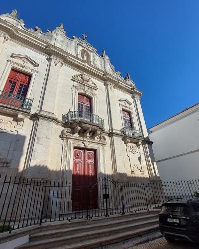 A low-angle photograph of a white stone church facade with red wooden doors and window shutters, featuring a black metal fence in the foreground and a black car parked on the street to the right.