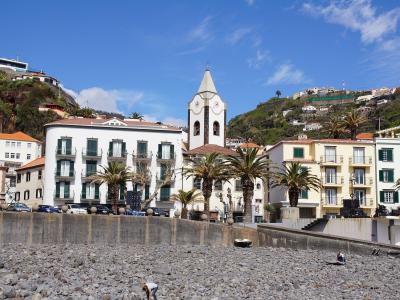 A pebble beach in the foreground with a row of white and yellow buildings, palm trees, and a church tower rising behind them on a sunny day.