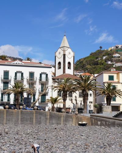 A pebble beach in the foreground with a row of white and yellow buildings, palm trees, and a church tower rising behind them on a sunny day.