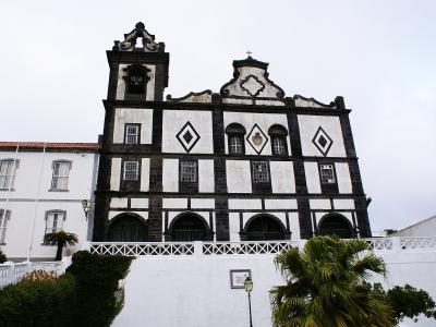 The image shows a church facade with distinctive black basalt stone framing and white plaster infill, featuring diamond-shaped motifs, arched ground-level openings, and a bell tower, situated in a coastal setting with palm trees and a white retain